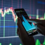 man holding a cellphone with stock chart in the baground